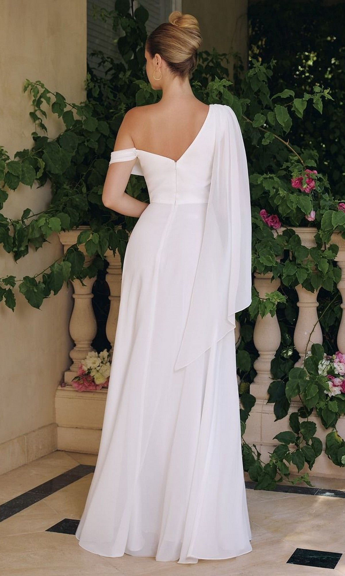 A woman wearing the Asymmetrical Off-White Bridal Gown: Amoris T716W, featuring a long chiffon skirt, stands with her back to the camera before a balustrade adorned with green vines and pink flowers.